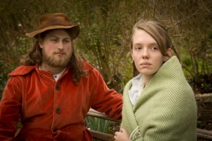 Man in russet jacket and soft hat siting on bench with girl wrapped in green shawl