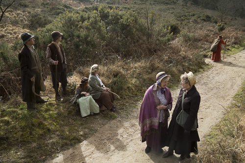 Two women in nineteenth century costume standing on a dirt track talking, with other people in the background